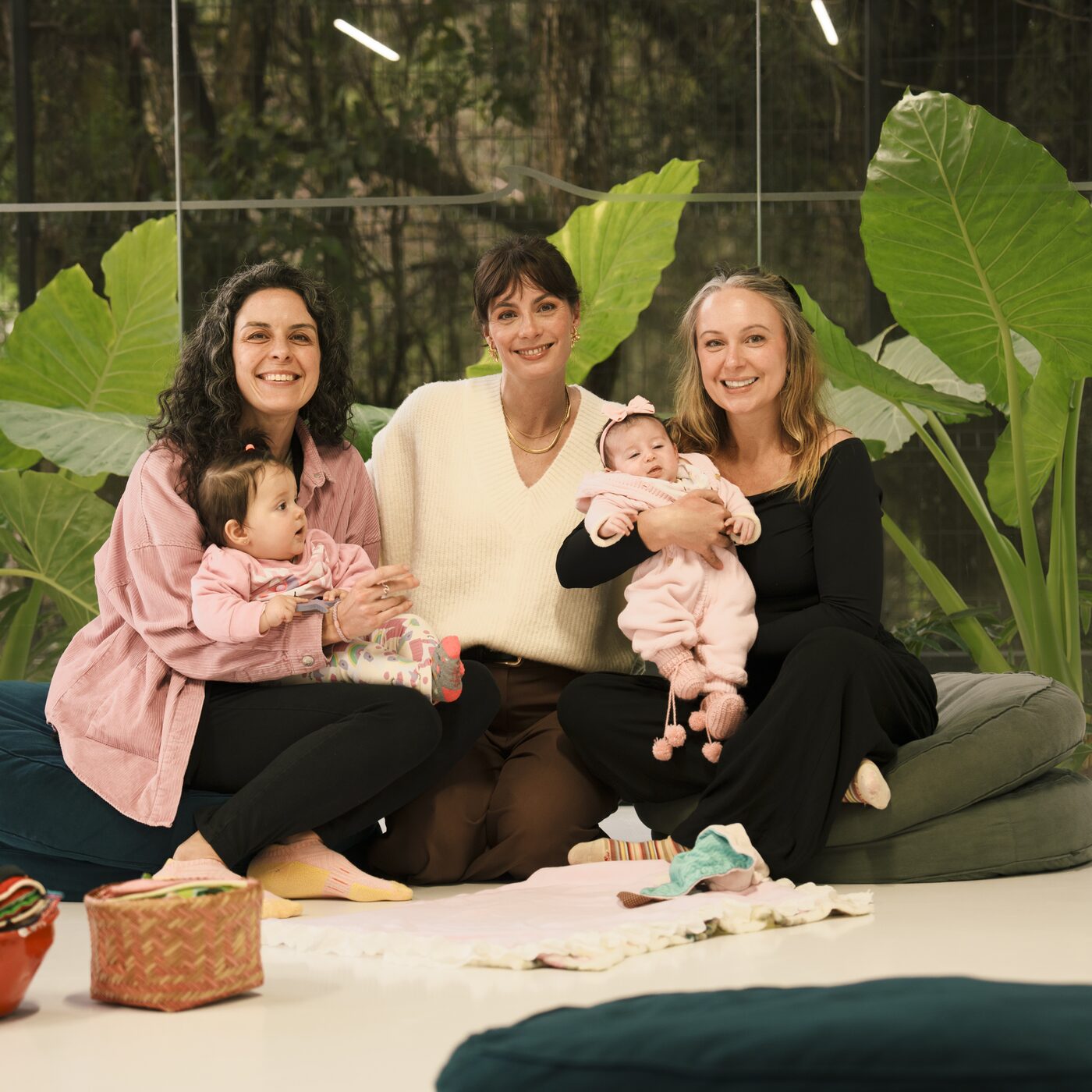 Três mães sorridentes com seus bebês no colo, sentadas juntas em um encontro do Parental Lab, com plantas tropicais ao fundo