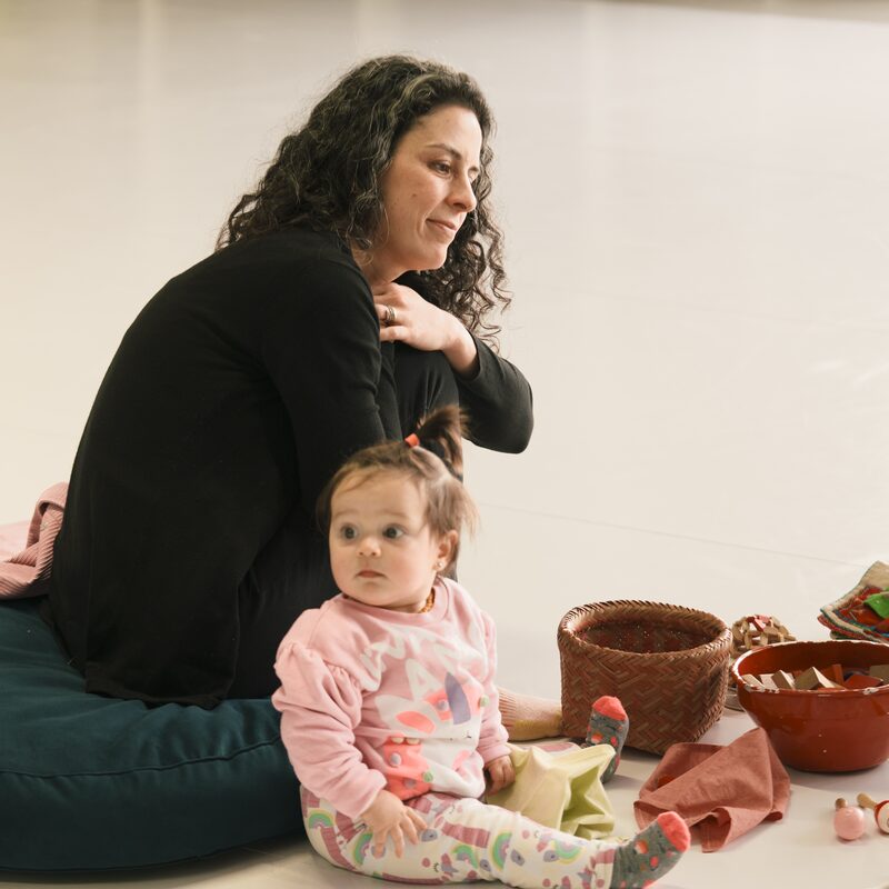 Mãe sentada no chão com a filha bebê, cercadas por cestos de brinquedos sensoriais do encontro