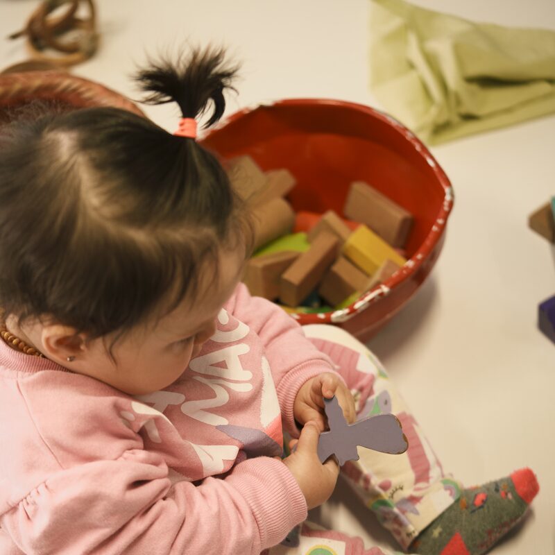 Bebê sentada no chão explorando uma tigela cheia de brinquedos de madeira durante o encontro