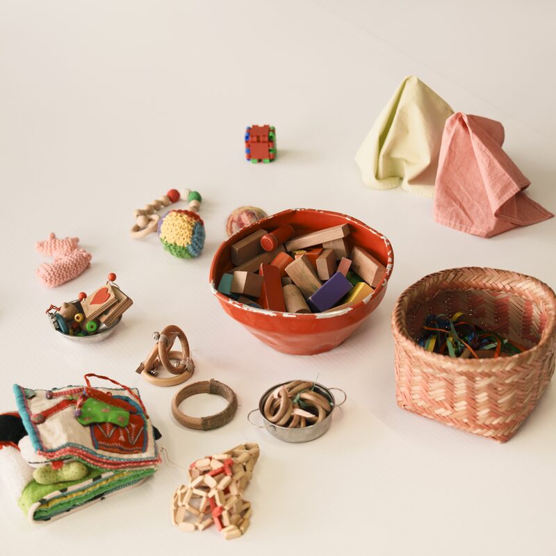 Brinquedos sensoriais de madeira, tecidos e chocalhos organizados no chão para a atividade com os bebês