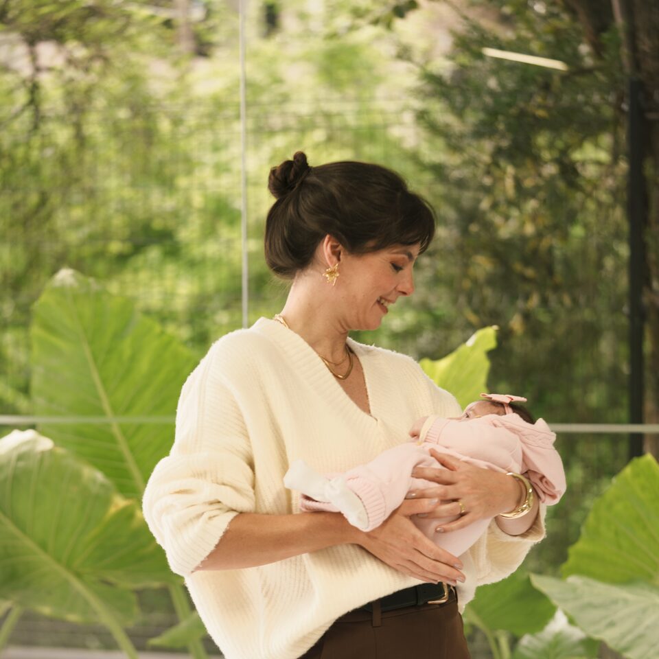 Luciana Lima em pé, segurando um bebê recém-nascido no colo, com vegetação exuberante ao fundo